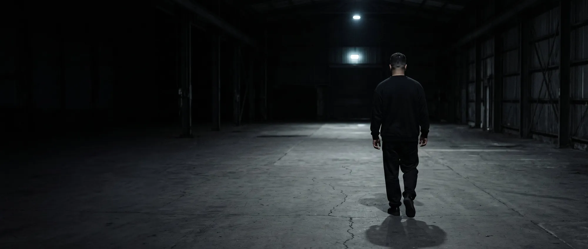 A man walking away through a vast dark interior toward a single distant light.