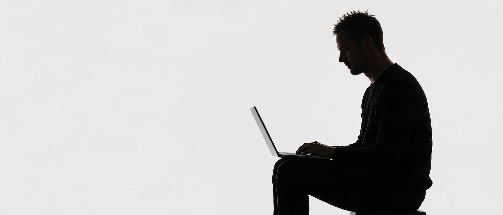 Silhouette seated, working on a laptop balanced on one knee.