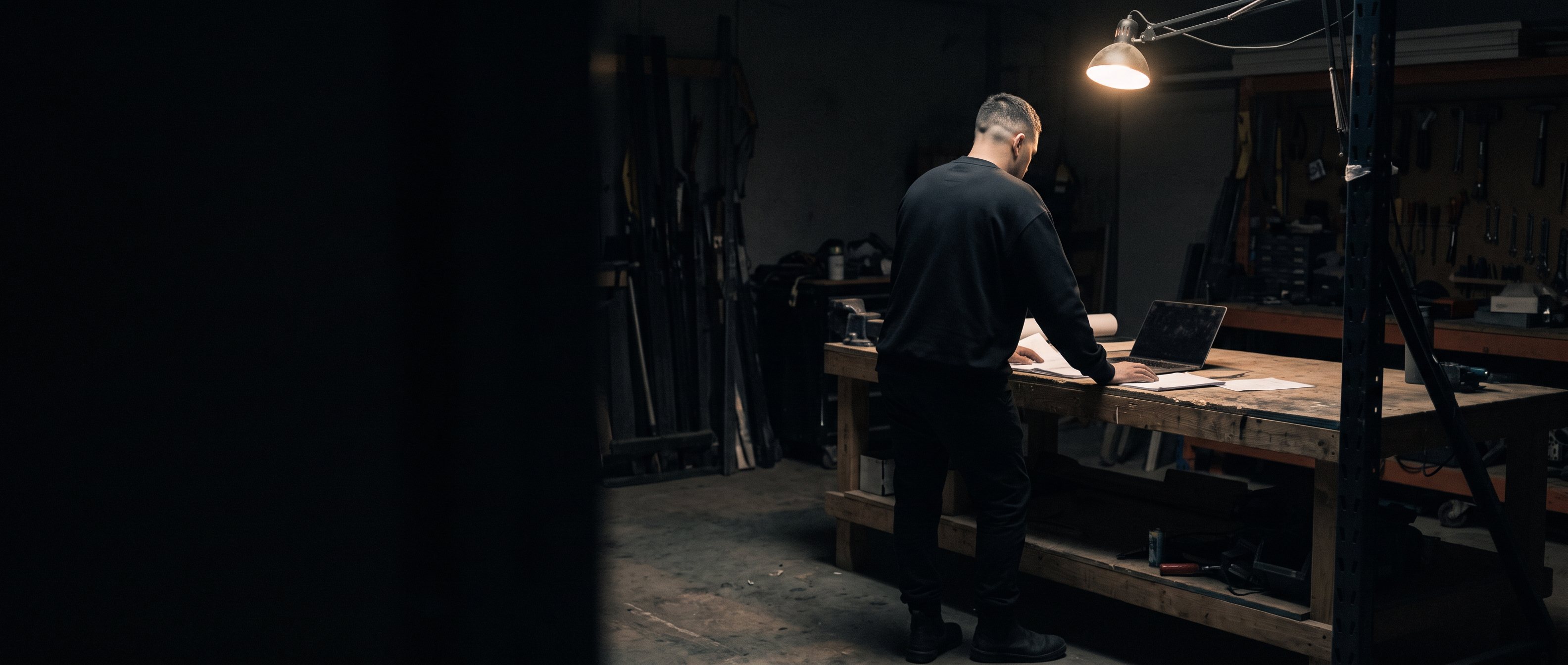A man in work clothes checking a tablet on a site after dark, back to camera.