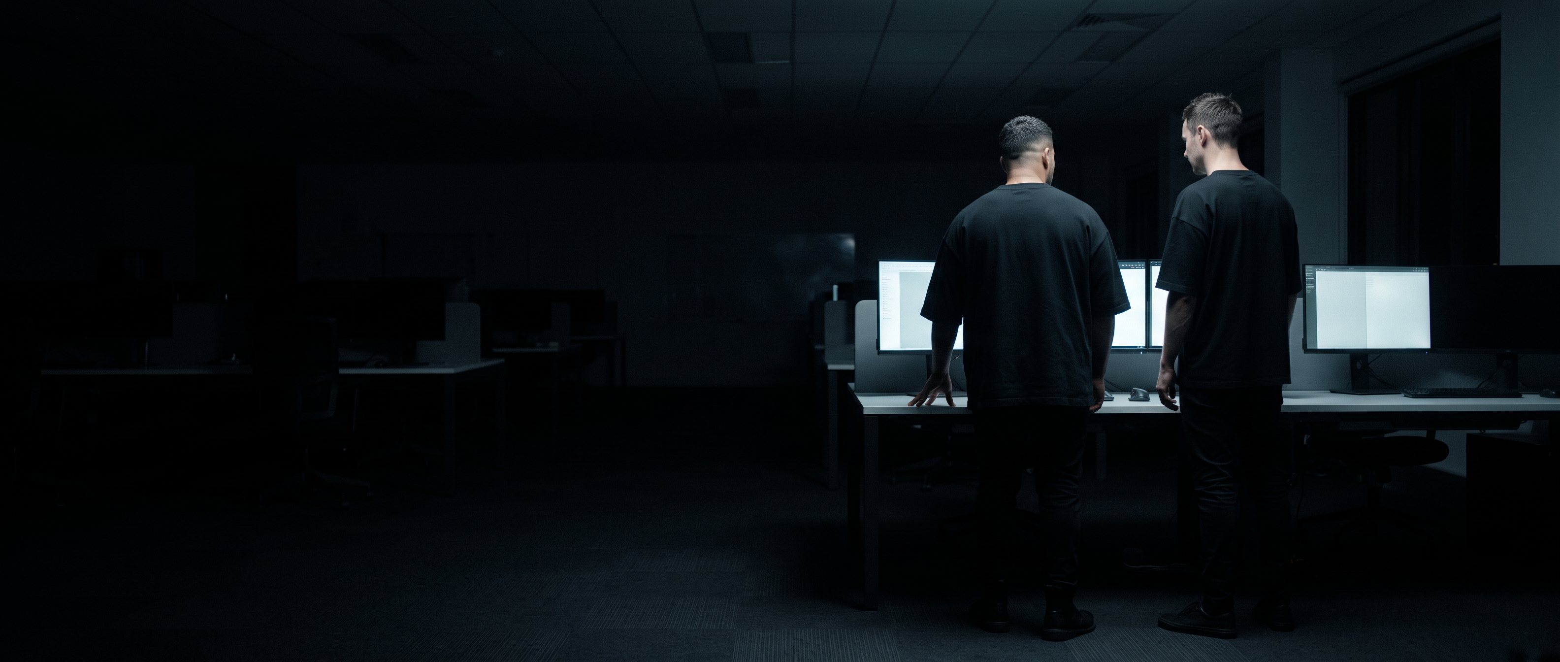 Two men at a desk in an unlit office at night, backs to camera, monitors lit.