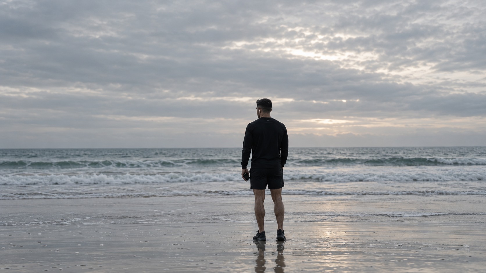 A man standing alone on the sand at dawn, looking out at the surf.
