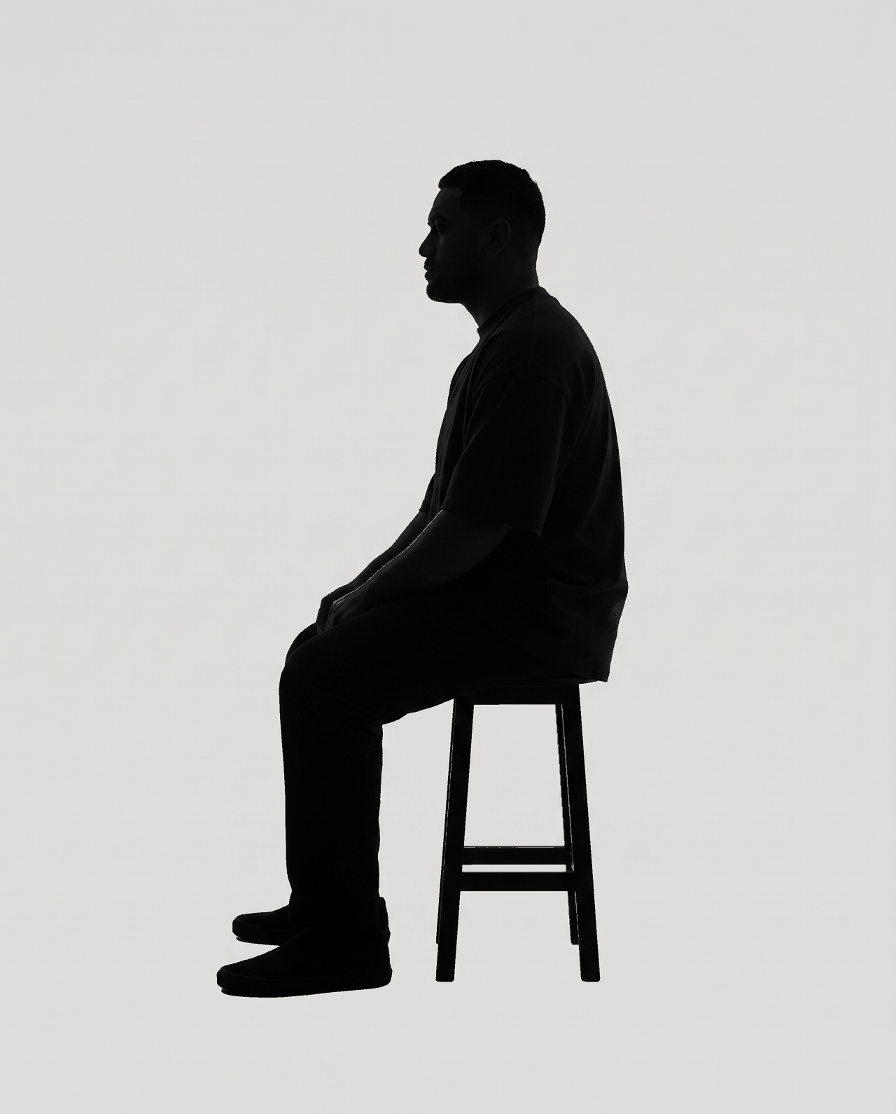 Dark silhouette of a man sitting on a stool in profile, hands in his lap, on flat grey.