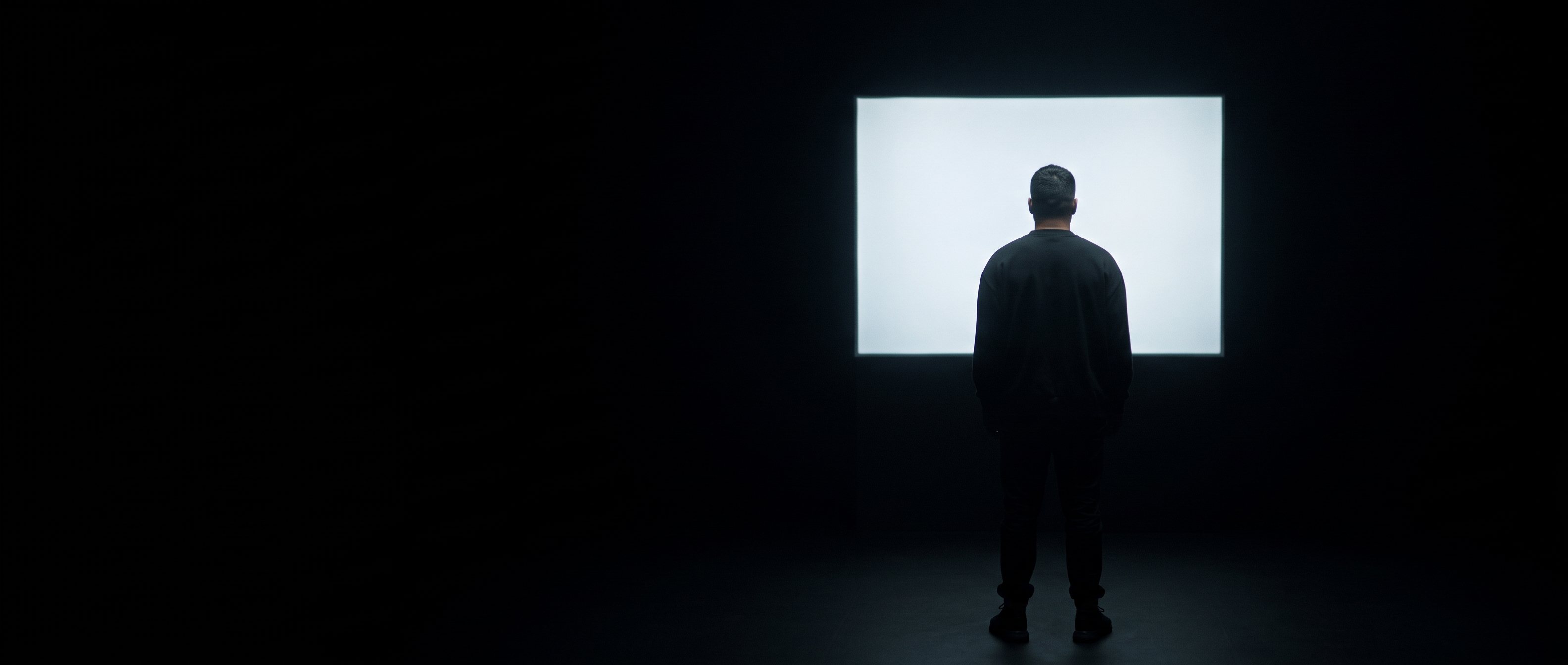 A man on a phone call in a dark room lit by one window, back to camera.