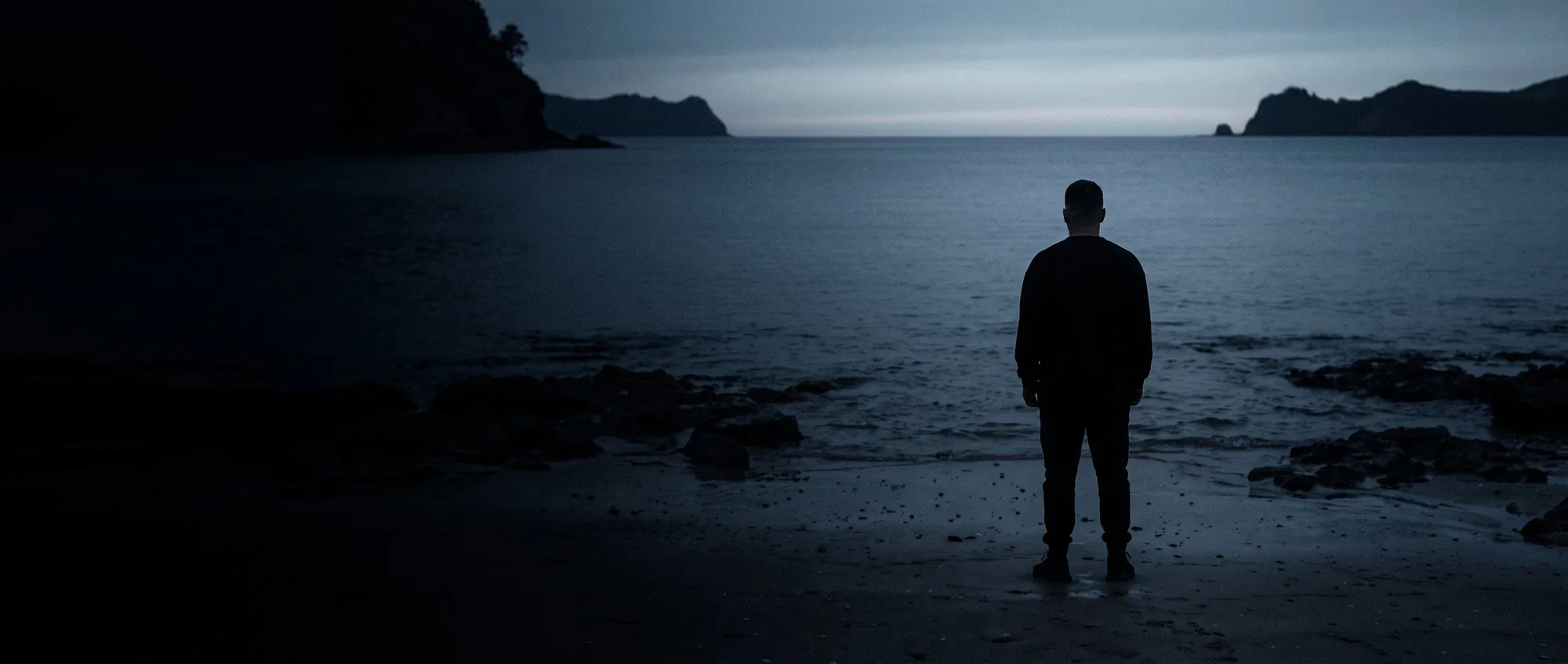 A man standing at the tideline of a dark coastline before sunrise, back to camera.